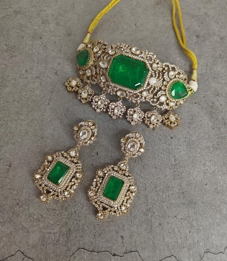 May include: A gold choker necklace with a large emerald green gemstone and smaller white gemstones. Matching earrings with emerald green gemstones and white gemstones.