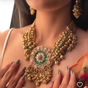 以下が含まれることがあります： 金色のネックレスとイヤリングのセット。ネックレスは、緑色の中心、赤いアクセント、複雑なディテールが施された大きな円形ペンダントが特徴です。セットには、ぶら下がっているビーズ要素が含まれており、フォーマルな場面に適しています。