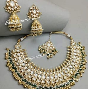 Pode incluir: Um colar de ouro com um grande pingente adornado com pedras brancas e contas verdes. O colar &eacute; combinado com brincos combinando com um design semelhante. Os brincos t&ecirc;m um charme pendente em forma de sino.