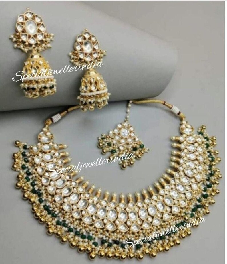 Pode incluir: Um colar de ouro com um grande pingente adornado com pedras brancas e contas verdes. O colar &eacute; combinado com brincos combinando com um design semelhante. Os brincos t&ecirc;m um charme pendente em forma de sino.