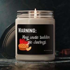 Puede incluir: Un tarro de vela blanco con una etiqueta negra que dice "ADVERTENCIA: Puede causar antojos repentinos de pastel." La etiqueta también presenta una manzana roja y una rebanada de pastel de manzana.