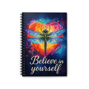 Può includere: Quaderno a spirale con una vivace illustrazione di una libellula con ali blu e turchese, su uno sfondo a forma di cuore nei toni dell'arancione, giallo e rosso. Le parole "Believe in yourself" sono scritte in elegante corsivo.