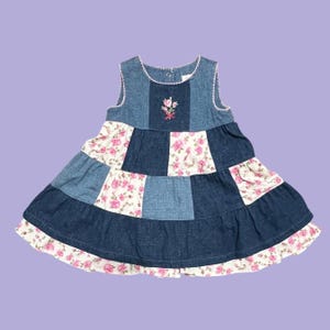May include: A sleeveless dress featuring a patchwork design of dark blue denim and white fabric with a pink rose floral pattern. The bodice includes a small embroidered rose detail at the center neckline, finished with white scalloped trim.