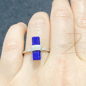 May include: A silver ring with a rectangular, deep blue gemstone and a white accent. The ring is set on a thin silver band. The design is modern and minimalist, with a contrast of colours and shapes. The gemstone is approximately 1 cm in length.
