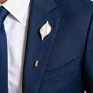 May include: A navy blue suit jacket worn with a crisp white collared shirt and a matching navy blue necktie. A white, leaf-shaped lapel pin with a metallic gold-toned edge is attached to the left side of the jacket, paired with a small gold pin.