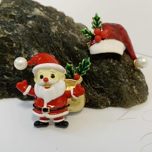 May include: Festive enamel pins with a Christmas theme, depicting a Santa Claus figure and a Santa hat. The Santa pin is red, white, and black, with a gold sack. The hat pin is red and white, with green holly and pearl accents.