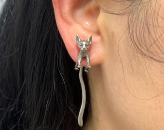 May include: A silver cat-shaped earring. The cat is depicted in a sitting position with a long, curved tail. The earring is attached to the ear, showcasing the detailed design of the feline figure.