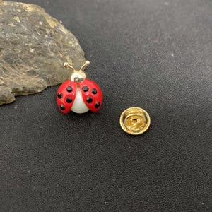 Puede incluir: Un broche de mariquita con un caparazón de esmalte rojo con puntos negros y un centro blanco nacarado. El broche tiene una cabeza de metal dorado con dos antenas y un cierre de embrague circular de metal dorado separado para fijación.