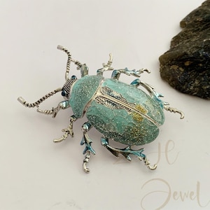 May include: A detailed, silver-toned beetle brooch with a turquoise and glittery body. The insect-themed jewelry piece has intricate leg and antenna details, and small blue accents. The brooch is set against a white background.