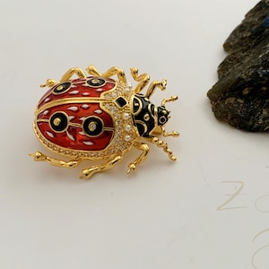 May include: A gold-coloured ladybird brooch with a red and black enamel design. The brooch is decorated with clear crystals and small pearls. The ladybird's body is red with black spots and a gold outline.