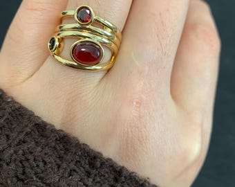 May include: A gold-toned ring with three bands, featuring a small round red stone, a small black stone, and a larger oval red stone. The ring is worn on a finger, showcasing the jewelry's design and color.