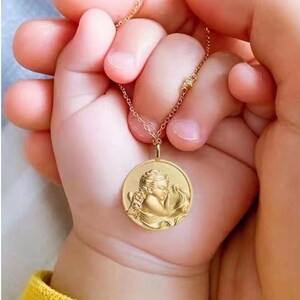 Pu&ograve; includere: Una collana con catena d'oro con un ciondolo rotondo che presenta una rappresentazione dettagliata di un angelo addormentato. Il ciondolo &egrave; tenuto da due mani, una delle quali &egrave; la mano di un bambino.