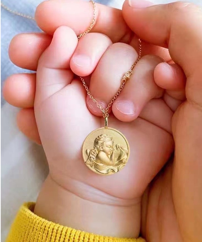Pu&ograve; includere: Una collana con catena d'oro con un ciondolo rotondo che presenta una rappresentazione dettagliata di un angelo addormentato. Il ciondolo &egrave; tenuto da due mani, una delle quali &egrave; la mano di un bambino.
