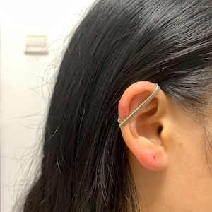 May include: A silver-colored ear cuff is worn on the upper ear. The jewelry is a simple, curved band that wraps around the ear's cartilage. The person has dark hair.
