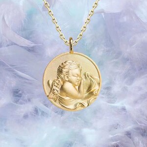 Pu&ograve; includere: Una collana d'oro con un ciondolo rotondo che presenta un cherubino addormentato con le ali. Il ciondolo &egrave; su uno sfondo di piume bianche.