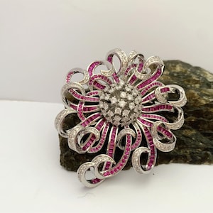 May include: A floral-shaped brooch with a silver-tone metal setting. The brooch features a central cluster of small, round, clear gemstones surrounded by curved ribbons of pink gemstones and clear gemstones. The brooch is set on a dark, textured surface.