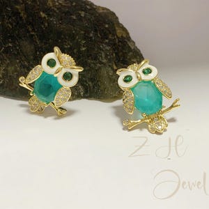 May include: Two gold-toned owl-shaped earrings. Each owl features a teal-colored gemstone body, white enamel accents, and sparkling crystal details. The owls are perched on a branch, with green eyes. The earrings are set against a white background.