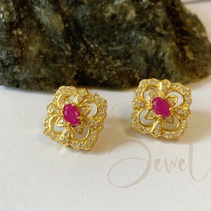 May include: A pair of gold-toned floral stud earrings. Each earring features a central oval-shaped ruby gemstone, surrounded by smaller clear gemstones. The earrings are set against a white surface.