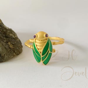 Könnte beinhalten: Goldfarbener Ring in Form einer Zikade mit strukturiertem Körper, grünen Emailleflügeln und lila Edelsteinaugen. Der Ring ist vor einem weißen Hintergrund mit einem Felsen links platziert.