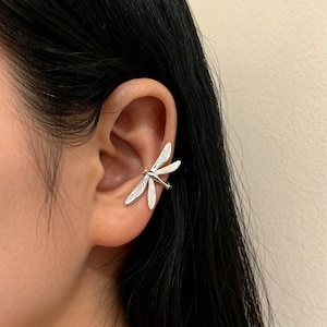 Puede incluir: Un brazalete de oreja de libélula plateada. El brazalete se envuelve alrededor de la oreja, con las alas de la libélula extendiéndose hacia afuera. Las alas tienen detalles grabados y el cuerpo de la libélula es liso.