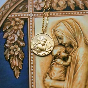 Pu&ograve; includere: Una collana d'oro con un ciondolo rotondo che presenta un'immagine dettagliata di un cherubino con le ali. Il ciondolo &egrave; appeso sopra un'immagine di una madre e un bambino.