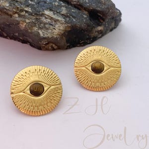 Pode incluir: Um par de brincos dourados. Cada brinco apresenta um design circular com uma pedra castanha central, assemelhando-se a um olho. Os brincos têm um padrão de raios de sol que irradia do centro. O texto "ZH Jewelry" é visível.