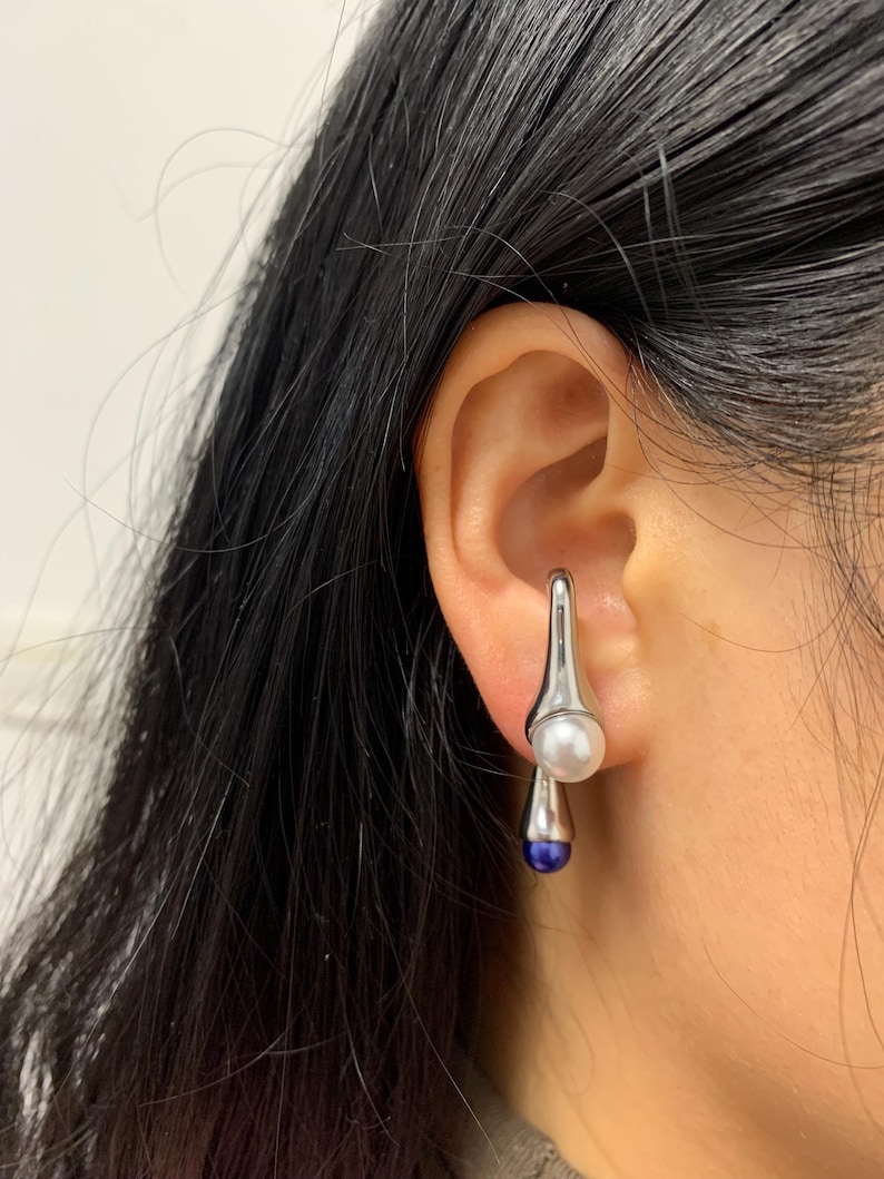 May include: Silver drop earrings with a white pearl and a blue gemstone. The earrings are dangling from the earlobe.