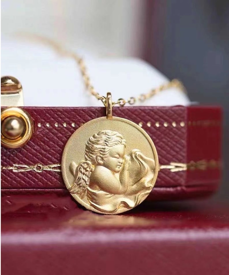 Pu&ograve; includere: Una collana d'oro con un ciondolo rotondo che presenta una scultura tridimensionale dettagliata di un cherubino con ali, che tiene un panno nelle sue mani.
