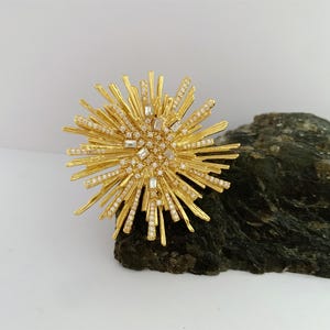Könnte beinhalten: Eine goldfarbene, sternförmige Brosche mit zahlreichen strahlenförmigen Spitzen. Die Brosche ist mit kleinen, klaren Kristallen und größeren, rechteckigen Kristallen in der Mitte verziert. Die Brosche liegt auf einem dunkelgrünen, strukturierten Felsen vor weißem Hintergrund.