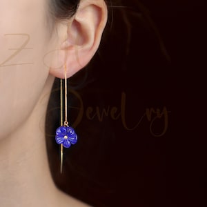 Puede incluir: Un par de pendientes de cadena dorada con un dije de flor azul. Los pendientes son largos y cuelgan del lóbulo de la oreja.