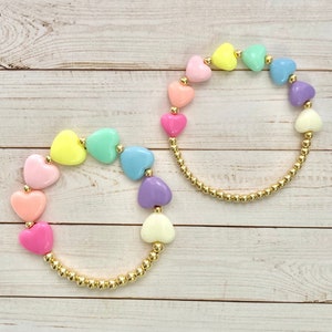 Puede incluir: Dos pulseras con cuentas doradas y cuentas en forma de coraz&oacute;n de color pastel. Las pulseras est&aacute;n sobre un fondo de madera blanco.
