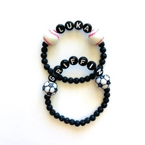 May include: Two black beaded bracelets with the names Luka and Griffin spelled out in white letters. The bracelets are decorated with black and white soccer balls and white baseballs.