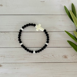 May include: A black and white beaded bracelet with a white cross charm. The bracelet is on a white wooden surface.
