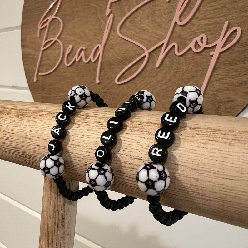 May include: Three black beaded bracelets with white soccer ball charms. Each bracelet spells out a name: "JACK", "OLIV", and "REED".