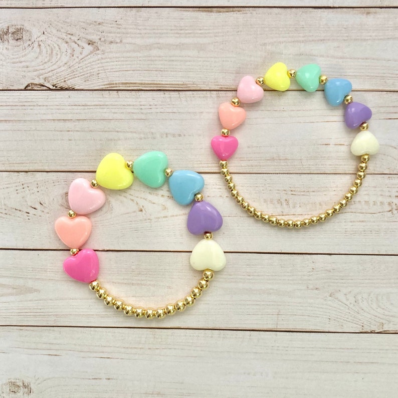 Puede incluir: Dos pulseras con cuentas doradas y cuentas en forma de coraz&oacute;n de color pastel. Las pulseras est&aacute;n sobre un fondo de madera blanco.