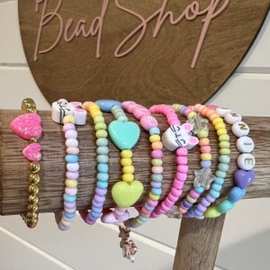 May include: A collection of colorful beaded bracelets with various charms, including hearts, stars, and animal faces. The bracelets are arranged on a wooden display stand.