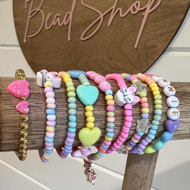 May include: A collection of colorful beaded bracelets with various charms, including hearts, stars, and animal faces. The bracelets are arranged on a wooden display stand.