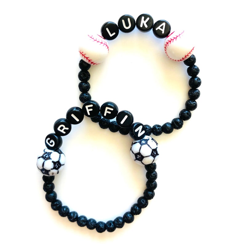 K&ouml;nnte beinhalten: Zwei schwarze Lava-Armb&auml;nder mit wei&szlig;en Buchstabenperlen, die die Namen "Luka" und "Griffin" buchstabieren. Jedes Armband hat einen wei&szlig;en und schwarzen Baseball- und Fu&szlig;ball-Anh&auml;nger.