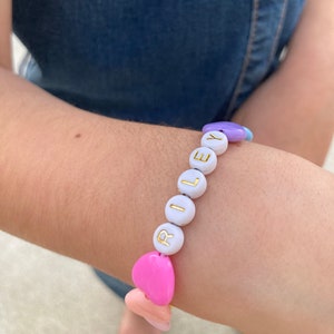 May include: A white beaded bracelet with the name "RILEY" spelled out in gold letters. The bracelet is accented with pink, purple, and blue heart-shaped beads.