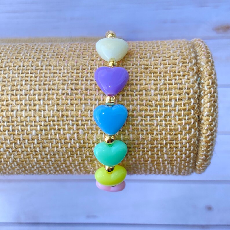 Puede incluir: Un brazalete de coraz&oacute;n arco&iacute;ris con cuentas de colores pastel. El brazalete est&aacute; hecho con cuentas de color amarillo claro, verde, azul, morado y blanco en forma de coraz&oacute;n. Las cuentas est&aacute;n enhebradas en una cadena dorada.