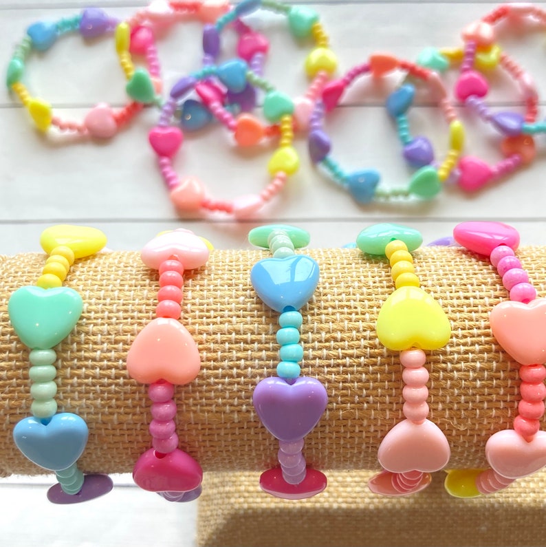 May include: A set of pastel-colored heart-shaped bracelets. The bracelets are made of plastic beads and are strung on elastic cord. The bracelets are arranged in a rainbow order, from yellow to purple.