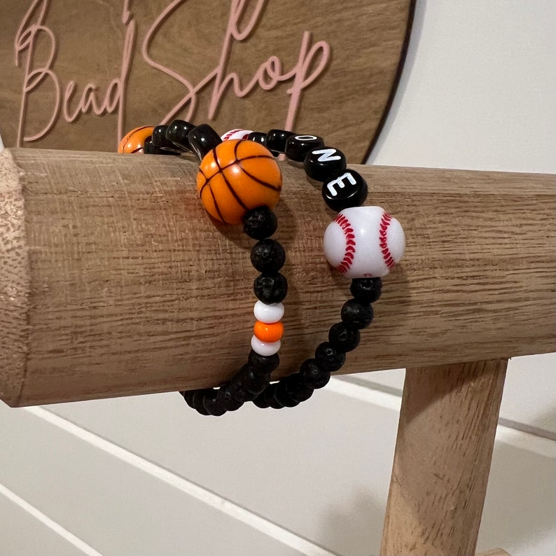 May include: Two beaded bracelets with black lava beads, orange and white beads, and sports-themed charms. One bracelet has a basketball charm and the other has a baseball charm. The bracelets spell out "ONE" in black letter beads.