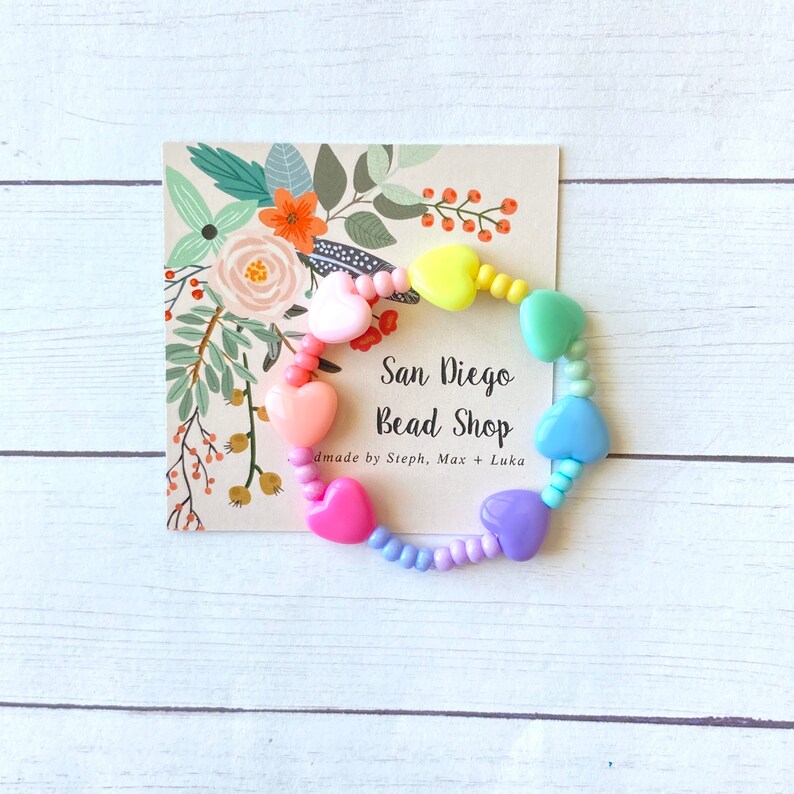 May include: Rainbow heart-shaped bead bracelet with pastel pink, yellow, green, blue, and purple beads. The bracelet is on a white card with the text "San Diego Bead Shop" and "made by Steph, Max + Luka".