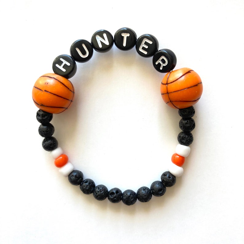 K&ouml;nnte beinhalten: Ein schwarzes und orangefarbenes Basketball-Armband mit dem Namen "HUNTER", der in wei&szlig;en Buchstaben auf schwarzen Perlen geschrieben ist.