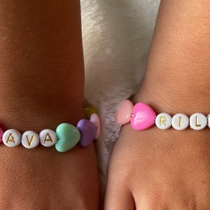 May include: Two bracelets with heart-shaped beads and white letter beads spelling out the names "AVA" and "RILEY". The bracelets are on the wrists of two people.
