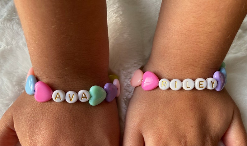 May include: Two bracelets with heart-shaped beads and white letter beads spelling out the names "AVA" and "RILEY". The bracelets are on the wrists of two people.
