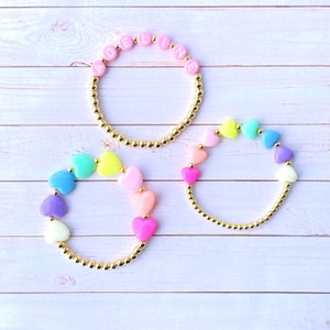 Puede incluir: Tres pulseras de oro con cuentas de colores. Una pulsera tiene el nombre "ADELINE" escrito en cuentas rosas. Las otras dos pulseras tienen cuentas en forma de coraz&oacute;n de arco&iacute;ris.