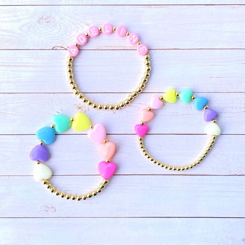 Puede incluir: Tres pulseras de oro con cuentas de colores. Una pulsera tiene el nombre "ADELINE" escrito en cuentas rosas. Las otras dos pulseras tienen cuentas en forma de coraz&oacute;n de arco&iacute;ris.