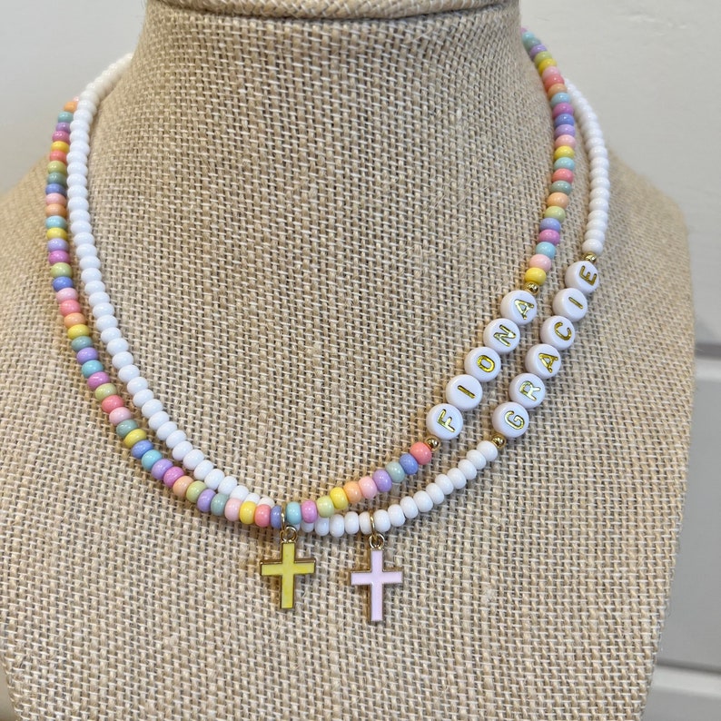 May include: A multi-colored beaded necklace with the names "Fiona" and "Gracie" spelled out in gold letters. The necklace has two small gold cross charms.