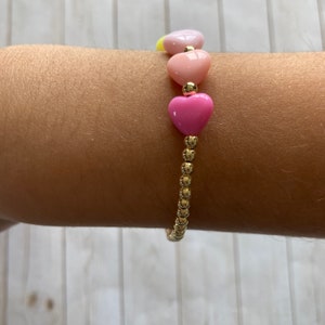 Puede incluir: Un brazalete dorado con una serie de cuentas en forma de coraz&oacute;n rosa y amarillo.